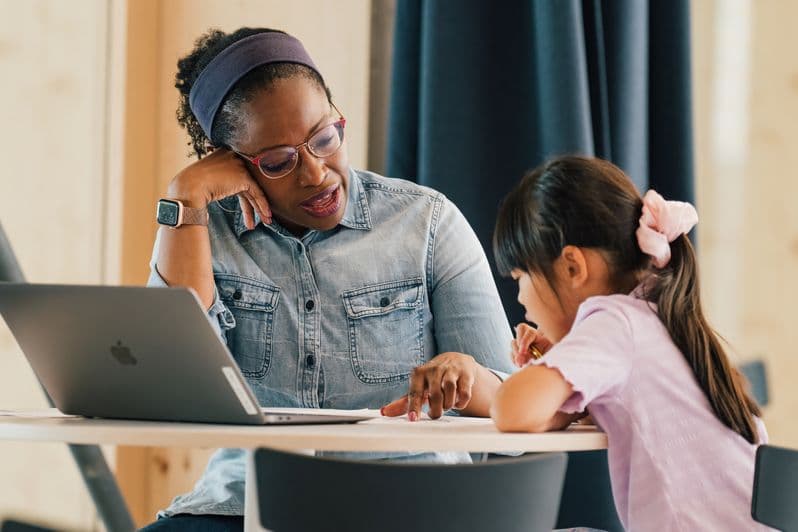 A woman in glasses helps a young girl with homework at a table with a laptop.